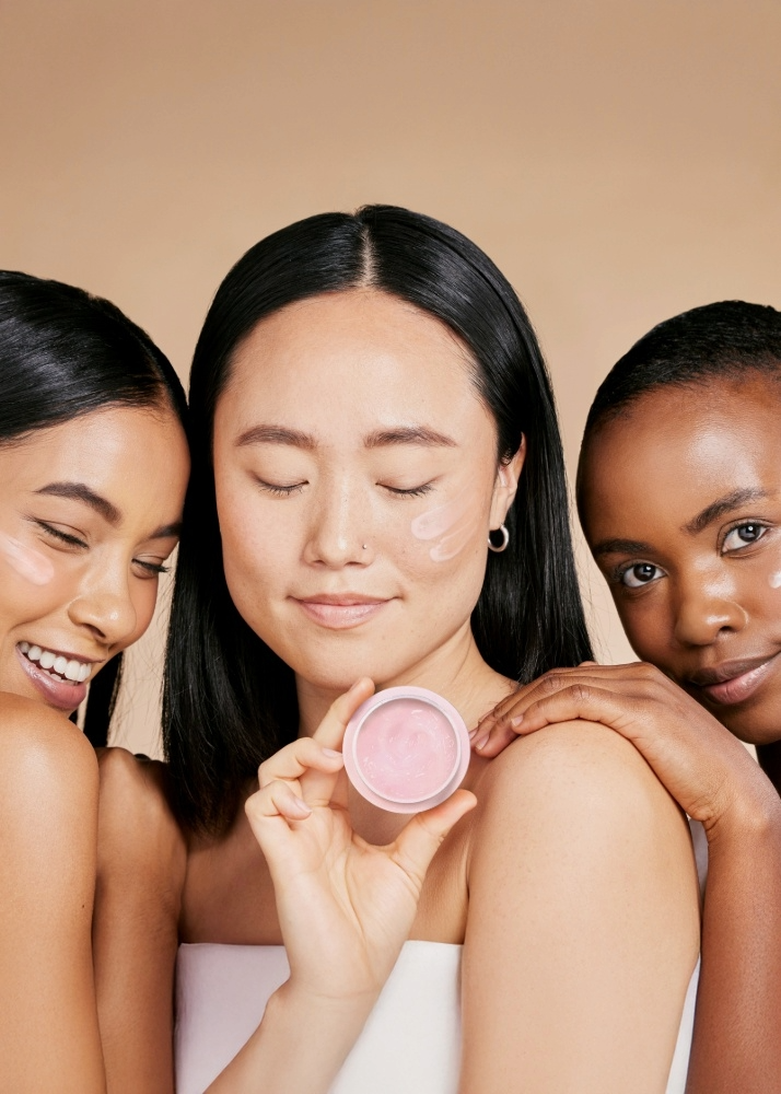 Three women holding a pink compact mirror against a beige background