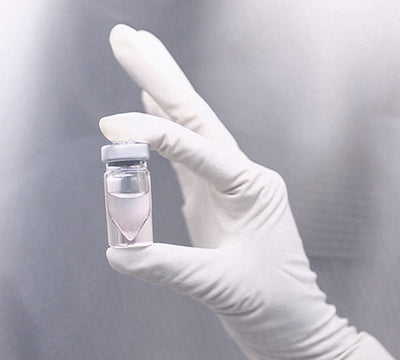 Hand wearing a white glove holding a small glass vial with a clear liquid against a gray background